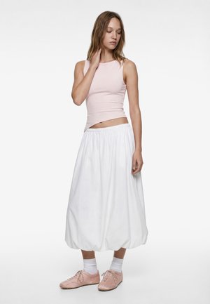 Jonge vrouw in een lichtroze mouwloze top, witte ballonvormige midirok, witte sokken en lichtroze veterschoenen, staand met een hand aan haar nek.