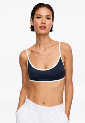 Frau mit kurzen dunklen Haaren, die ein marineblaues Crop-Top mit weißem Rand und weiße, hochgeschnittene Hosen vor weißem Hintergrund trägt.
