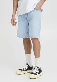 Pantalones cortos de mezclilla azul claro con un ajuste relajado, que presentan una cinturilla estándar y bolsillos. Combinados con zapatillas negras y calcetines blancos.