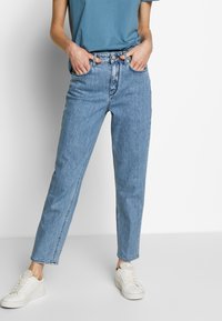 Vaqueros de tiro alto en azul claro, con corte recto, cinco bolsillos y trabillas para cinturón, combinados con una camiseta de cuello redondo azul y zapatillas blancas.