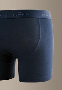 Boxeri scurți de culoare albastru închis cu cusături vizibile și o bandă de talie cu inscripția „NEXT SIGNATURE” pe un fundal neutru.