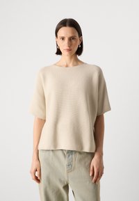 Beige strikket topp med korte ermer, rund halsutskjæring og løs passform; kombinert med lyseblå denim bukser. Teksturert stoffdetalj er synlig.