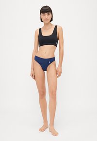 Črn športni nedrček in modri bikini hlački. Nedrček ima kvadratni izrez in široke naramnice, medtem ko hlačke vsebujejo majhen logotip in klasičen kroj.