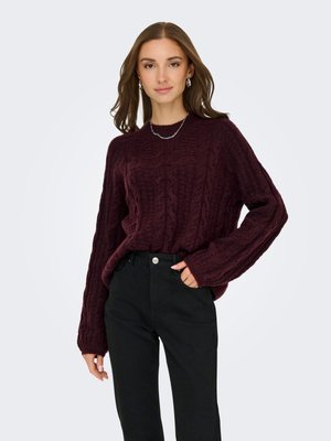 Vrouw die een donkerpaarse kabelgebreide trui draagt, zwarte hoge taille broek, zilveren ketting en zilveren oorringen, poserend met één hand in de zak.