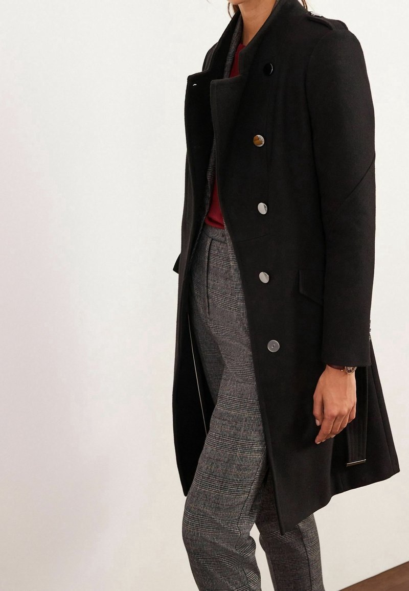 Personne portant un manteau noir croisé, un haut bordeaux et un pantalon gris à carreaux, debout devant un fond blanc uni.