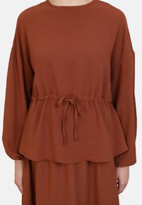Rust-colored top with long sleeves, cinched waist featuring a drawstring, and a textured fabric; scalloped hemline adds detail.