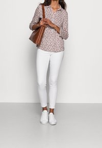 Kvinde iført hvid mønstret bluse, hvide skinny jeans, hvide sneakers og bærende en brun læder skuldertaske, som står op ad en hvid væg.