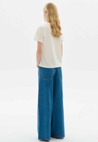 Blonde woman stands facing away wearing a white short-sleeve top and wide-leg blue denim jeans against a plain background.