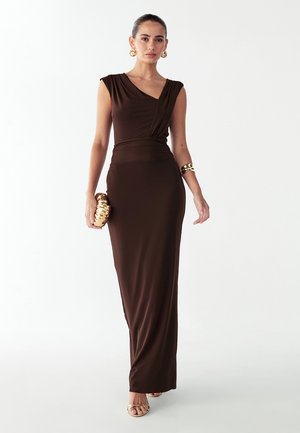 Bruine mouwloze maxi-jurk met een geplooide bovenkant en een aansluitend silhouet. Het model houdt een gouden clutch vast en draagt bijpassende gouden sieraden.