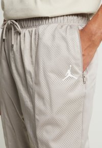 Person, der hellgraue Basketballshorts mit Mesh-Einsätzen, einer Reißverschlusstasche und einem weißen Jumpman-Logo trägt, mit der Hand in der Tasche.