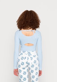 Camisola de mangas longas em azul claro com nervuras, com um recorte nas costas e detalhe em tiras, combinada com calças brancas com padrões de flores azuis.