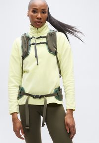 Ανοιχτό κίτρινο φλις πουλόβερ με ψηλό γιακά και πράσινες λεπτομέρειες. Διαθέτει δύο τσέπες στο στήθος και καφέ ρυθμιζόμενη ζώνη με αγκράφες.