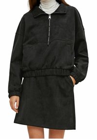 Vrouw in een zwart suède jurk met lange mouwen, een rits aan de voorkant en een elastische tailleband, gecombineerd met een witte coltrui eronder.