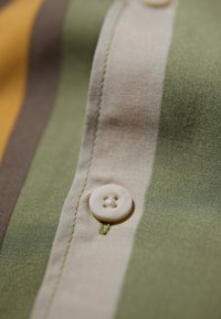Camisa de botones con rayas verdes, beige, marrón y amarillas. Tejido texturizado con un botón de cuatro agujeros en la costura del cuello.