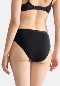 Mustat korkeavyötäröiset alushousut, valmistettu pehmeästä kankaasta. Niissä on sileät reunat ja yksinkertainen muotoilu, vähän ompeleita ja saumaton ulkonäkö.