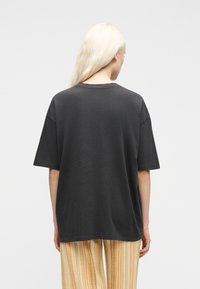 Άτομο με μακριά ξανθά μαλλιά που φοράει μια πολύ φαρδιά σκούρα γκρι κοντομάνικη μπλούζα και παντελόνι με κίτρινες-πορτοκαλί ρίγες, γυρισμένο προς τα πίσω σε λευκό φόντο.