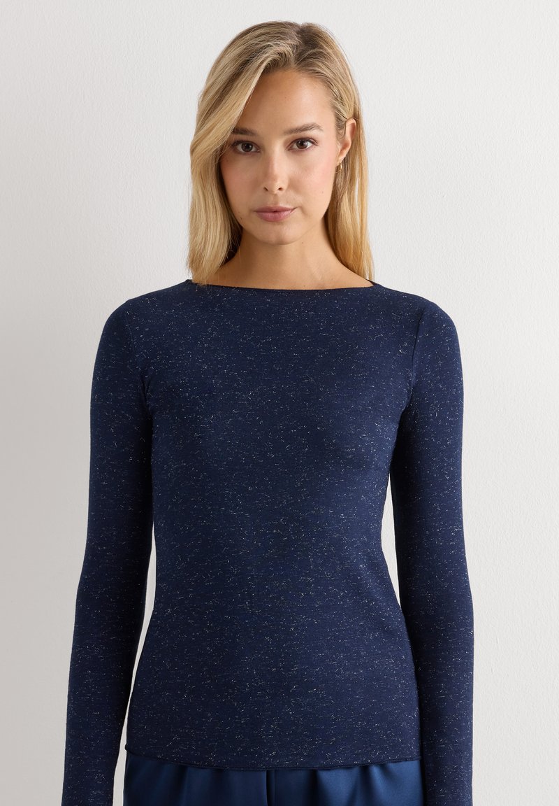 Vrouw met steil blond haar draagt een nauwsluitend marineblauw gestippeld shirt met lange mouwen tegen een effen lichte achtergrond.