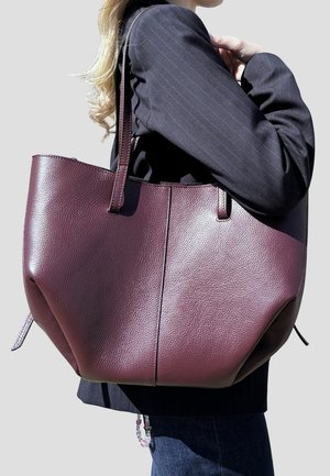 Donna con blazer nero che porta una grande borsa tote in pelle di colore viola scuro e dalla trama evidente sulla spalla, su sfondo semplice.