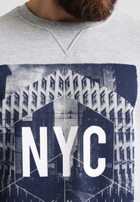 Sudadera gris con un gran gráfico azul marino del skyline de la ciudad de Nueva York, con "NYC" destacado en letras blancas en negrita.