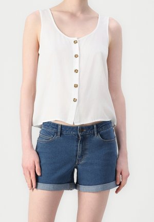 Kvinde iført en hvid, ærmeløs knapbluse og denimshorts med opkrogede kanter, stående mod en ensartet lys baggrund.