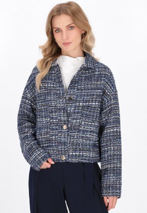 Giacca in tweed blu e bianco con una forma rettangolare, caratterizzata da un colletto, tre bottoni e una superficie strutturata.