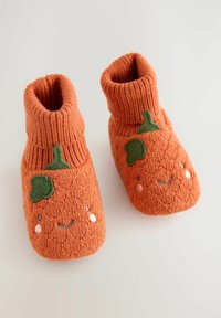 Des chaussons en tricot orange présentent un revers côtelé, un visage souriant et des accents de feuille verte. La finition texturée apporte chaleur et confort.