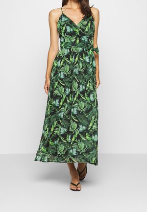 Femme portant une robe portefeuille sans manches à imprimé feuilles vertes avec une ceinture nouée à la taille, assortie à des tongs noires, debout devant un fond uni.