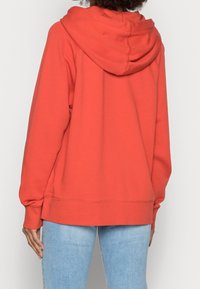 Felpa color arancio corallo con cappuccio ampio, vestibilità ampia, maniche lunghe e orlo a coste. Il tessuto morbido e testurizzato è visibile dalla vista posteriore.