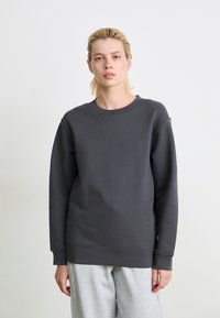 Felpa grigia scura con girocollo, polsini e orlo a costine, realizzata in tessuto liscio. Indossata con pantaloni della tuta grigi chiari, design minimal.