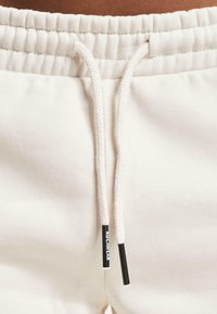 White sweatpants feature a drawstring waist with textured elastic, accented by black aglets and a logo tag. Soft, smooth fabric with a simple design.