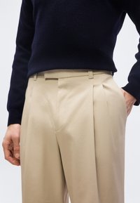 Personne portant un pantalon plissé beige et un pull noir à manches longues, une main dans la poche et l'autre détendue le long du corps.