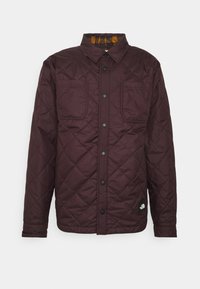 Burgundská prošívaná bunda se zapínáním na patentky, dvěma náprsními kapsami a kostkovanou podšívkou, vyrobená z odolné tkaniny. Černá logo nášivka na spodním lemu.