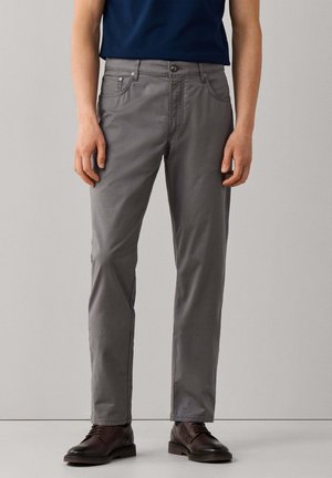 Man draagt grijze slim-fit broek, donkerbruine leren schoenen en een marineblauw overhemd, staand tegen een effen lichte muur.