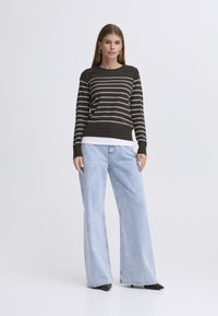 Temno črtast pulover z dolgimi rokavi, kombiniran s svetlo modrimi širokimi kavbojkami in črnimi škornji z ostrim prstom; model stoji pred enobarvnim ozadjem.