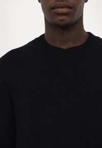 Pull en tricot bleu marine avec un motif de losanges texturé, col rond et poignets côtelés. Vue en gros plan du tissu du vêtement.
