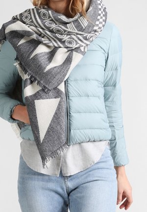Femme portant une veste matelassée bleu clair, une écharpe à motifs gris et blanc, une chemise gris clair, et un jean bleu clair, avec une main dans la poche de la veste.
