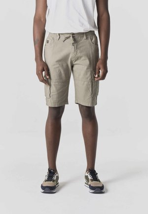 Mand iført beige cargoshorts, hvid t-shirt og grønne, sorte og beige sneakers, stående foran en neutral baggrund.