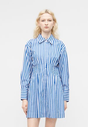 Γυναίκα με ξανθά μαλλιά που φοράει φόρεμα με μακριά μανίκια και γιακά, με κάθετες μπλε και λευκές ρίγες, στέκεται μπροστά σε απλό φόντο.