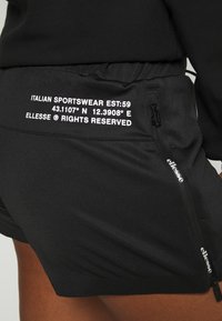 Svarta sportshorts med en zipficka. Har vit text på midjebandet: "ITALIAN SPORTSWEAR EST:59" och "ELLESSE® RÄTTIGHETER FÖRBEHÅLLNA."