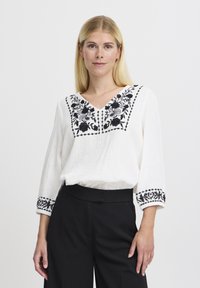 Blusa bianca con ricami floreali neri sul colletto e sulle maniche, con scollo a V e un tessuto leggero e strutturato.