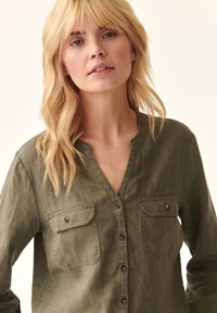 Camicia verde oliva con colletto a V, due tasche sul petto e tessuto testurizzato. Presenta bottoni marroni e una vestibilità rilassata.