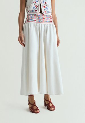 Mujer con falda larga blanca y chaleco corto con bordado floral rojo y azul, combinado con sandalias rojas tejidas con tacón.