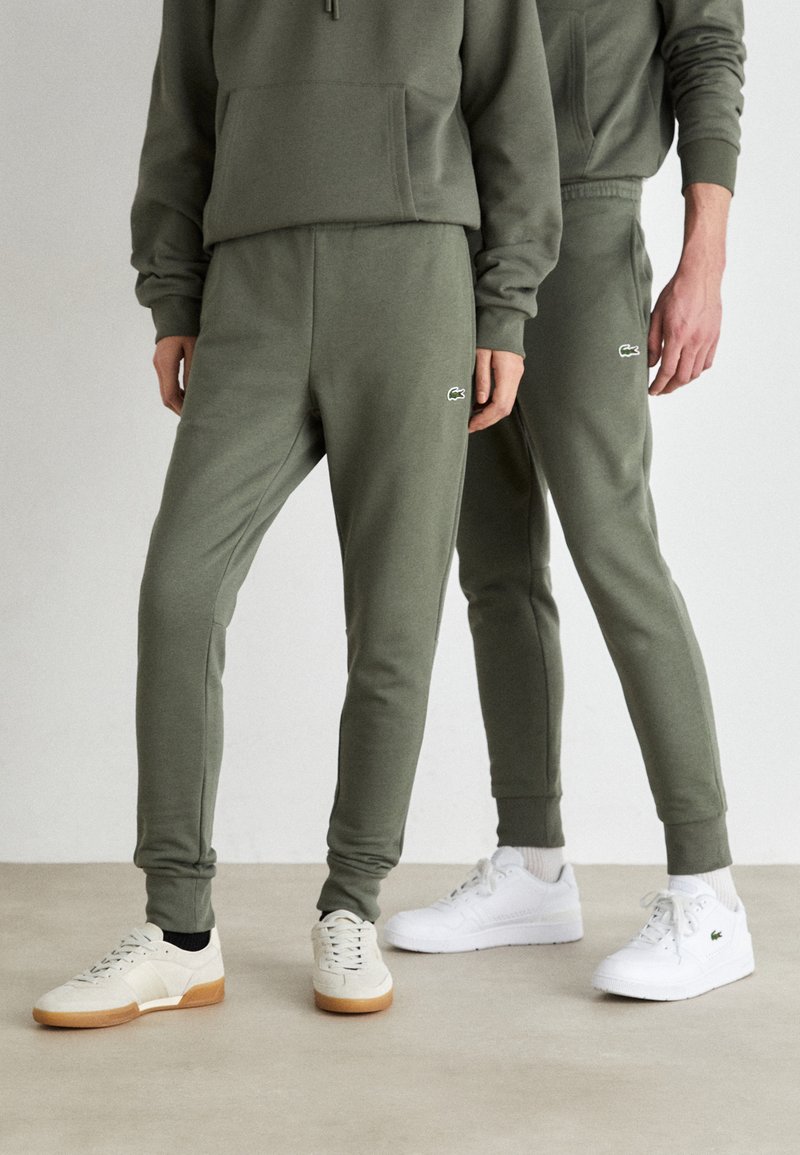 Olivgrüne Sweatpants und Hoodies aus weichem Stoff, mit gerippten Bündchen und kleinen Logo-Akzenten. Ein Paar weiße und kremfarbene Sneaker.