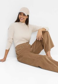 Pull en ribbed beige à manches longues associé à un pantalon large en velours côtelé marron, complété par une casquette beige avec un accent bleu.