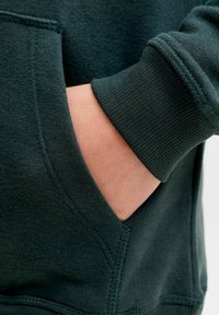 Sudadera de color verde oscuro con un bolsillo lateral. Cuenta con puños acanalados y una tela suave y texturizada. Los detalles de las costuras son visibles a lo largo de las uniones.