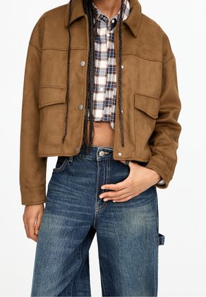 Person trägt eine braune kurze Jacke über einem karierten Crop-Hemd, kombiniert mit hoch taillierten, weit geschnittenen blauen Jeans, Hand auf der Taille.