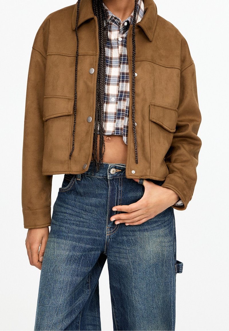Personne portant une veste courte marron sur une chemise courte à carreaux, associée à un jean bleu taille haute à jambes larges, main posée sur la taille.