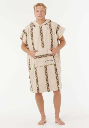 Joven de pie descalzo, llevando un poncho toalla beige con capucha, con rayas verticales marrones y un bolsillo frontal con el logo de Rip Curl.