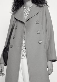 Manteau en laine grise au design croisé, avec de grands boutons, un large col et des poches latérales. Porté par-dessus un haut fleuri.