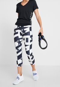 Kobieta w czarnej koszulce z krótkim rękawem i czarno-białych legginsach w wzory, trzymająca w jednej ręce zwoju opasek oporowych, ubrana w białe buty sportowe.